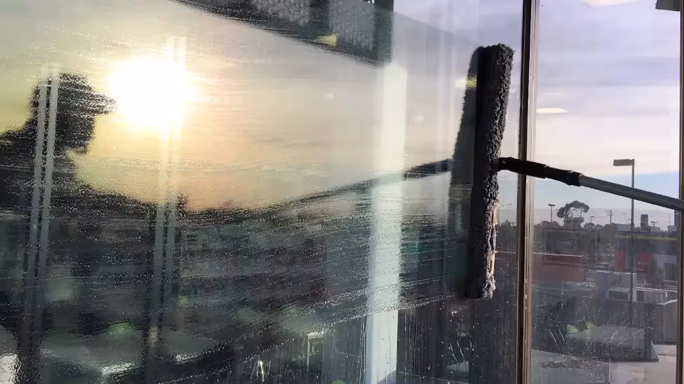 Close-up of a person using a long-handled washer to clean an exterior glass window at sunset.