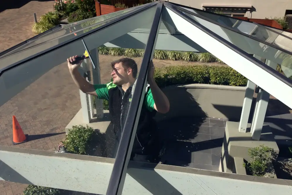 Worker in Bax Clean uniform, cleaning the glass roof of a modern building structure.