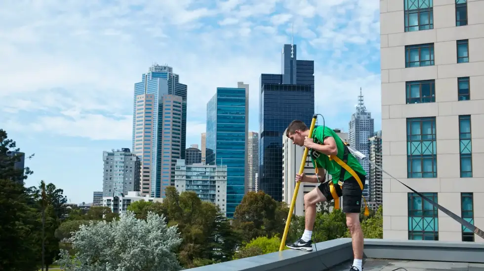 A worker wearing safety gear and a green uniform uses a long pole while standing on a rooftop with a city skyline in the background.