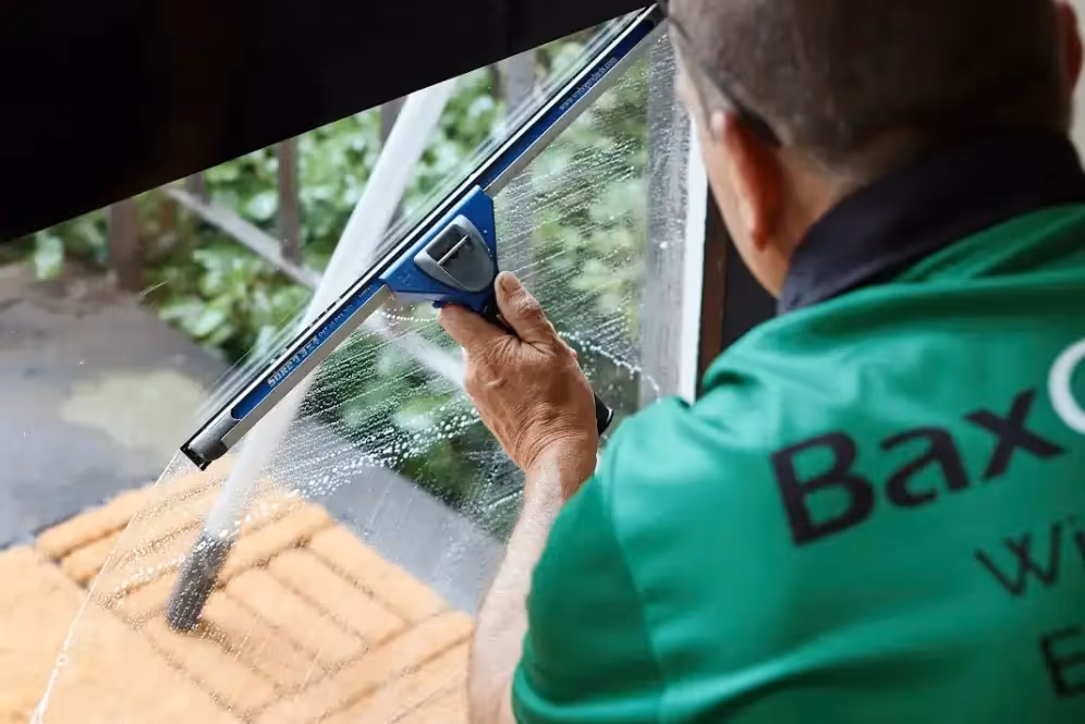 A professional window cleaner wearing a green Bax Clean uniform uses a squeegee to clean a glass door from the inside.