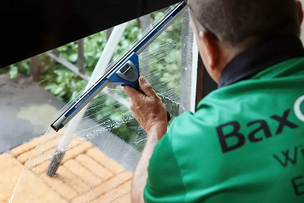 A professional window cleaner wearing a green Bax Clean uniform uses a squeegee to clean a glass door from the inside.