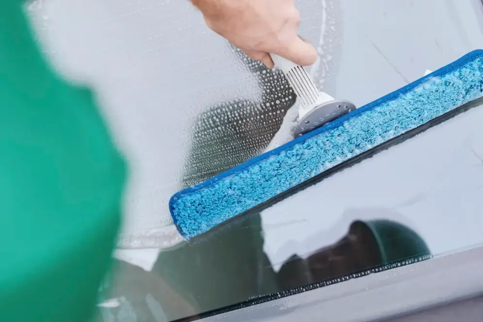 A Bax Clean worker uses a blue squeegee to clean a glass window, wearing a green uniform and brown work boots.