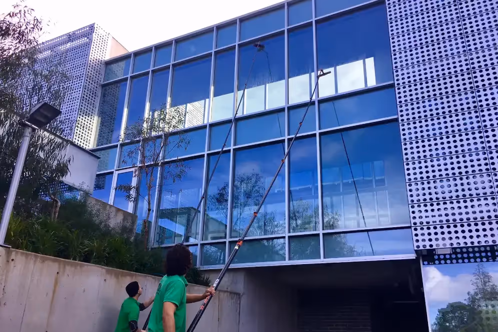 Two workers in green shirts cleaning large glass windows of a multi-storey building.