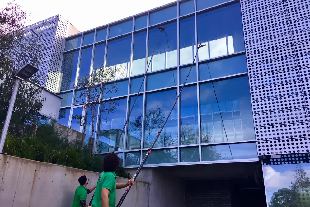 Two workers in green shirts cleaning large glass windows of a multi-storey building.