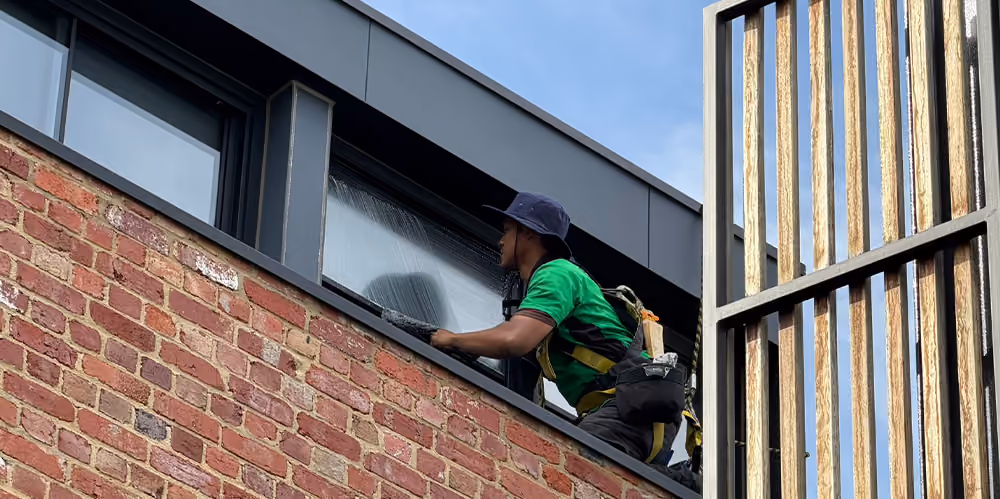 Technician cleaning upper-level windows beside timber privacy screens at the West Melbourne residential complex.