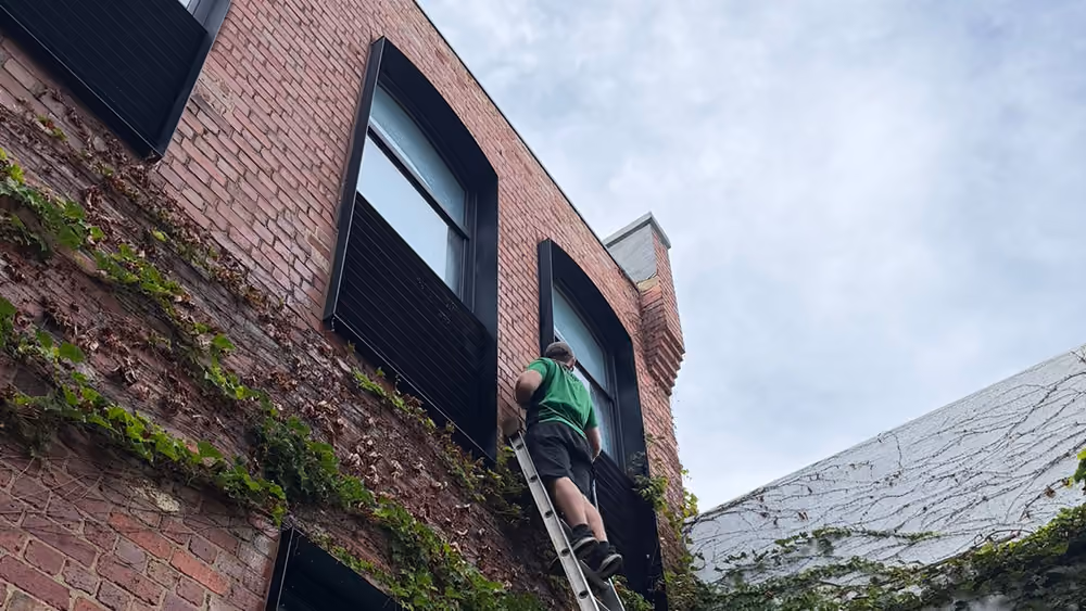 Bax Clean technician using a ladder to clean windows on a brick building