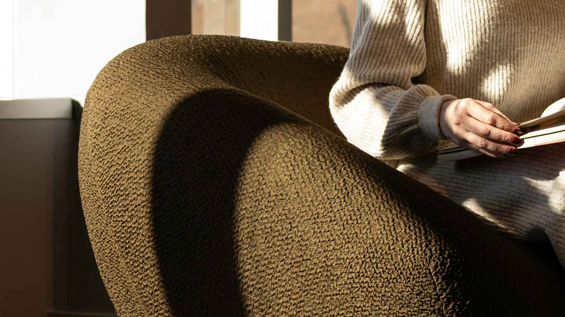 Close-up of a curved olive-green upholstered chair beside a person holding an open book.