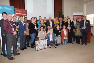 A group of people posing for a photo in front of a larege screen with a projection on it and multiple pull-up banners for different organisations.