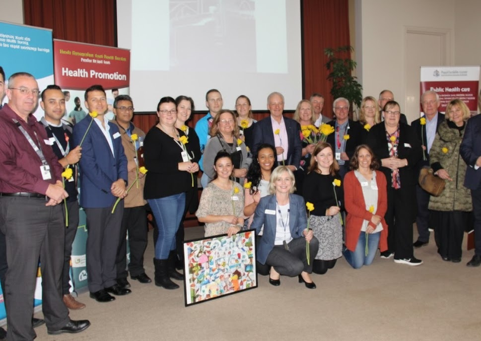 A group of people posing for a photo in front of a larege screen with a projection on it and multiple pull-up banners for different organisations.