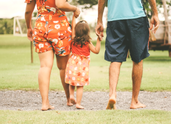 Little girl holding hands with a woman on the left and a man on the right. They are walking in a park away from camera.