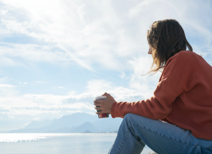 Woman sitting down with elbows on knees wearing a red sweater and jeans holding a coffee cup looking out at the ocean
