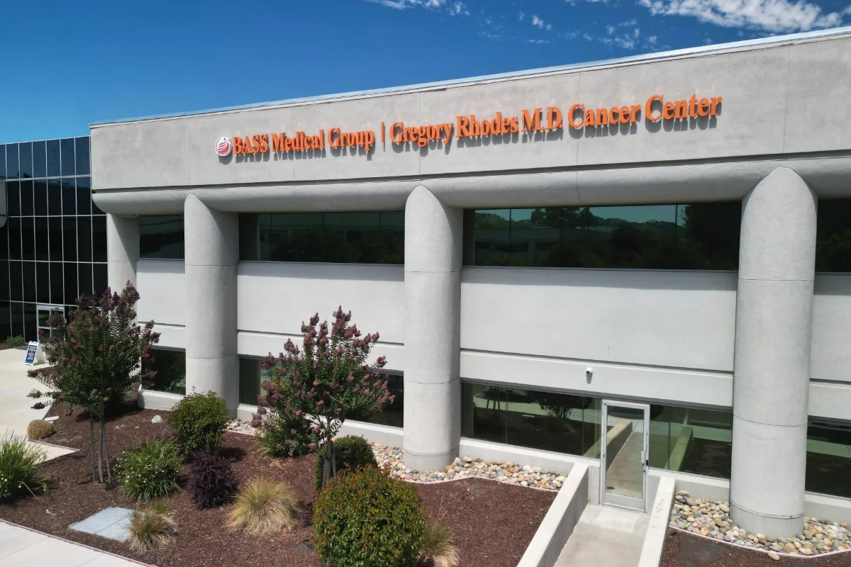 Exterior of the Gregory Rhodes MD Cancer Center, part of BASS Medical Group in Walnut Creek, featuring the off-white two-story building with the orange BASS logo and signage reading “BASS Medical Group | Gregory Rhodes MD Cancer Center,” surrounded by trees and landscaping.