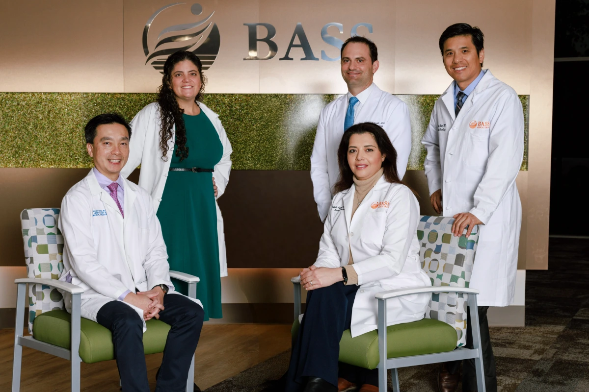 Dr. Danielle Bertoni, Dr. James Mitchell, and Dr. Kenneth Chao stand behind Dr. Tyler Kang and Dr. Soheila Azghadi, seated in front of a BASS Medical Group sign at Gregory Rhodes MD Cancer Center.