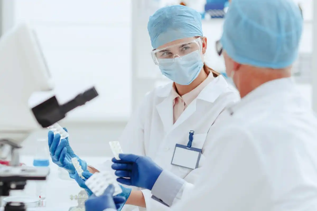 Two medical professionals in scrubs and masks reviewing data on a tablet while holding medication samples.