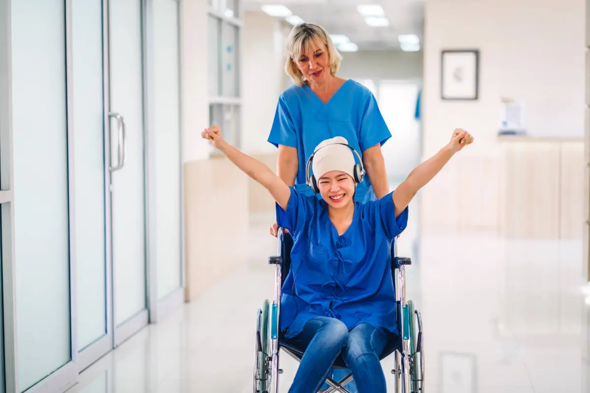 Cancer patient wearing a head wrap and headphones raises her fists in triumph while a nurse wheels her out of the Gregory Rhodes MD Cancer Center, symbolizing strength and recovery.