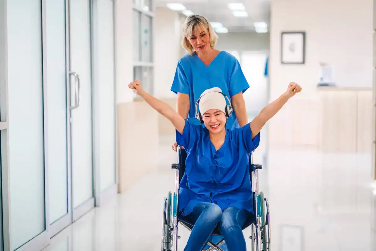 Cancer patient wearing a head wrap and headphones raising her fists while being wheeled by a nurse.