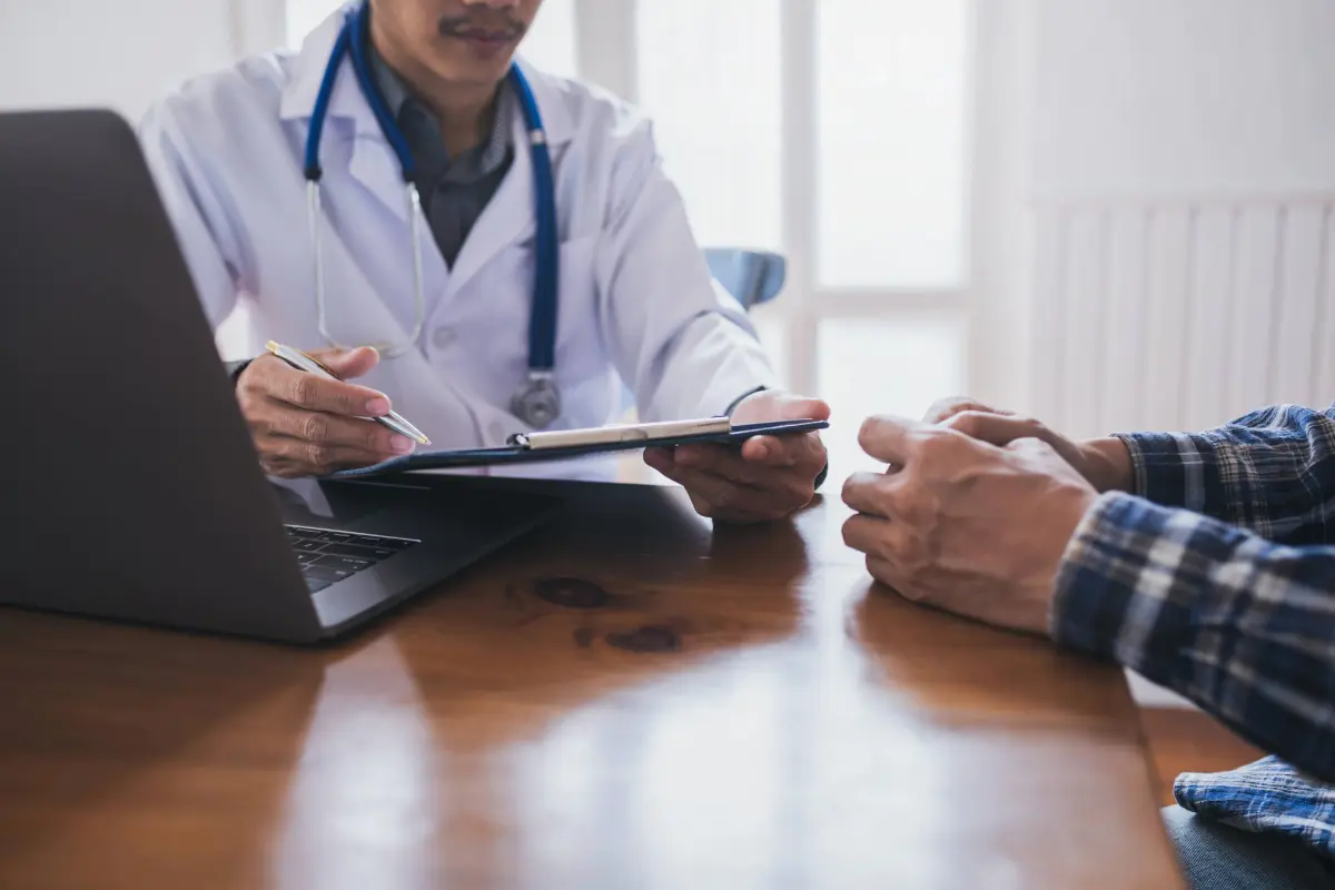 Male urologist in a white lab coat reviewing a medical chart with a male patient in a blue plaid shirt at Gregory Rhodes MD Cancer Center in Walnut Creek, symbolizing trusted prostate cancer care.