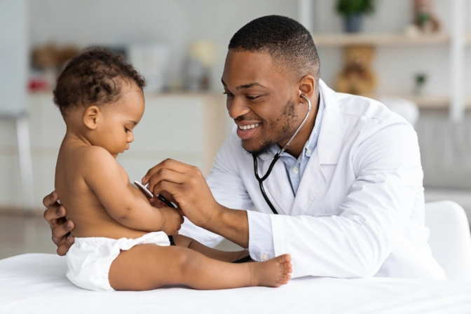 Pediatrics doctor using a stethoscope to check a baby's lungs while the baby curiously stares at the doctor.