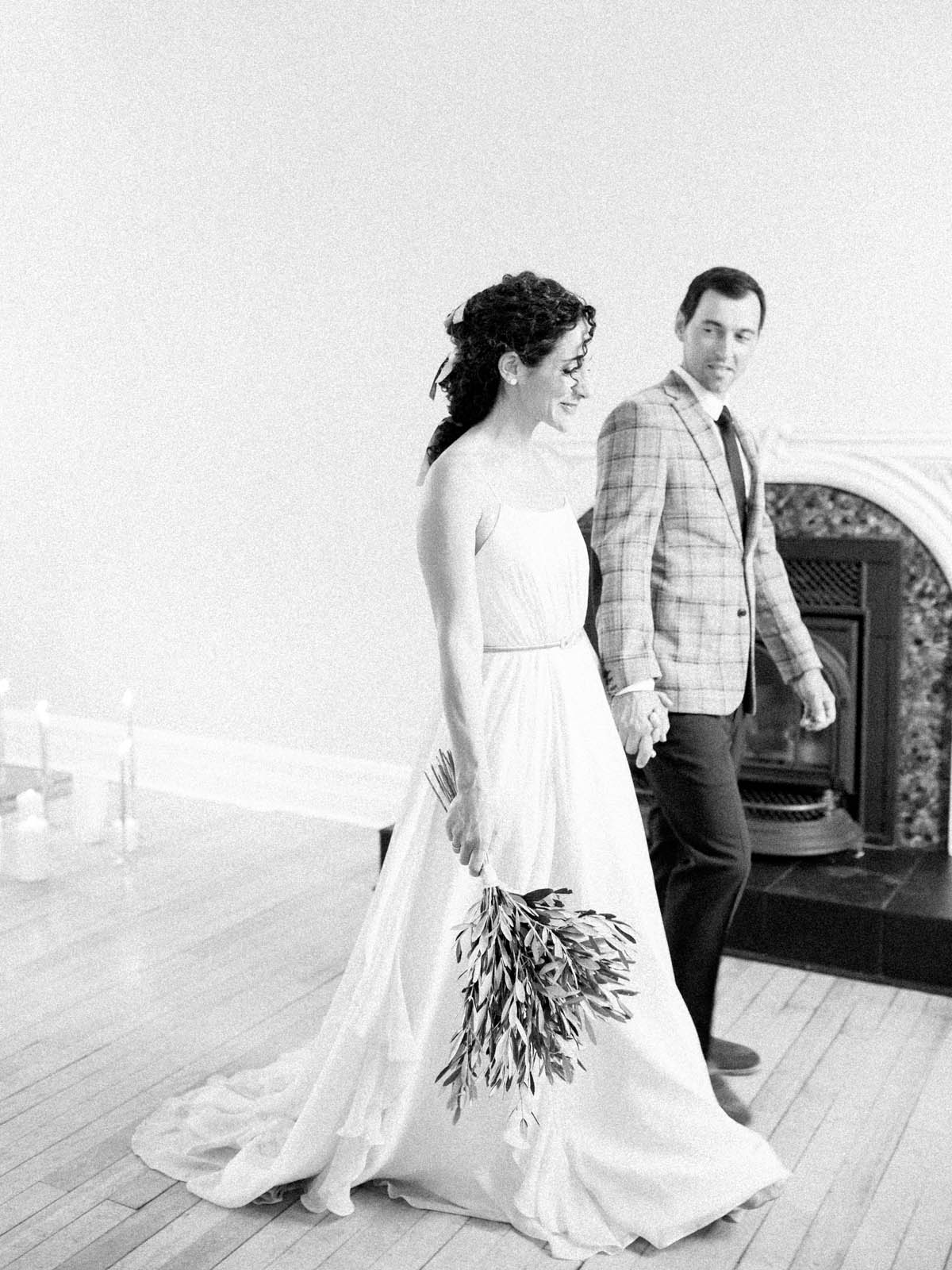 Bride carrying an olive branch bouquet as the couple walk past the fireplace