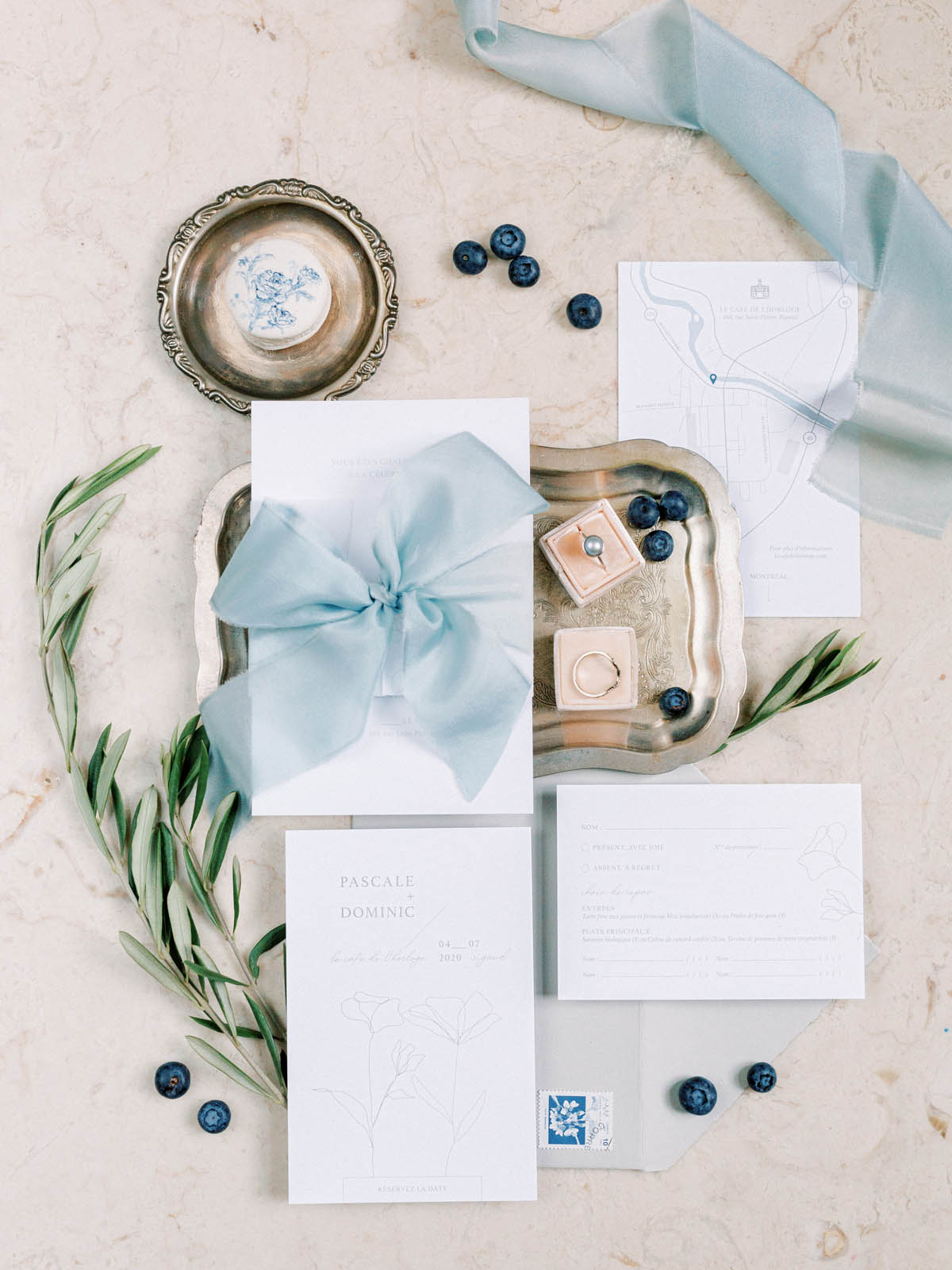 Invitation suite flat lay with blue silk ribbon, rings and olive branches