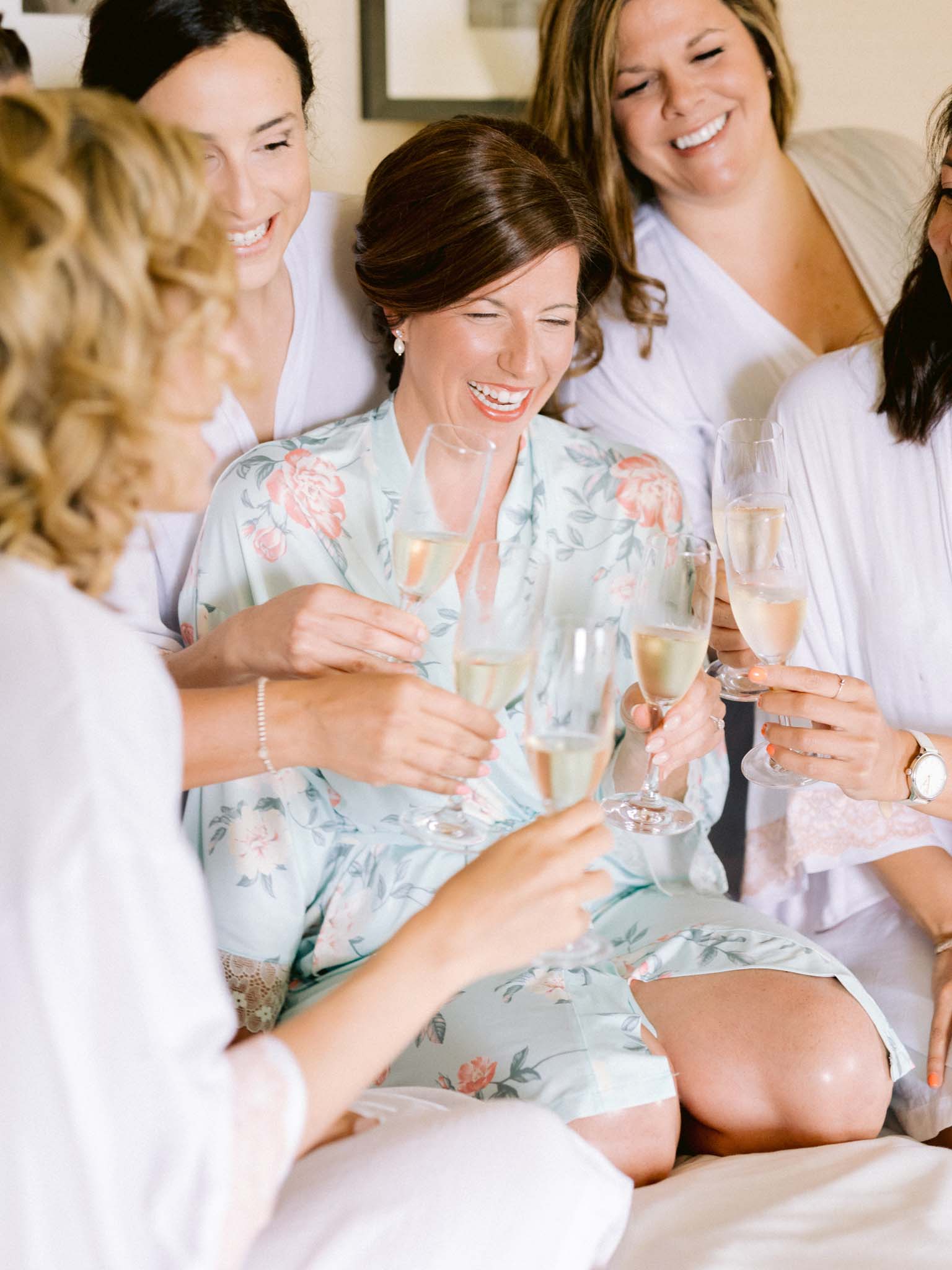 Bride laughing with her bridesmaids over champagne in floral robes on the morning of the wedding