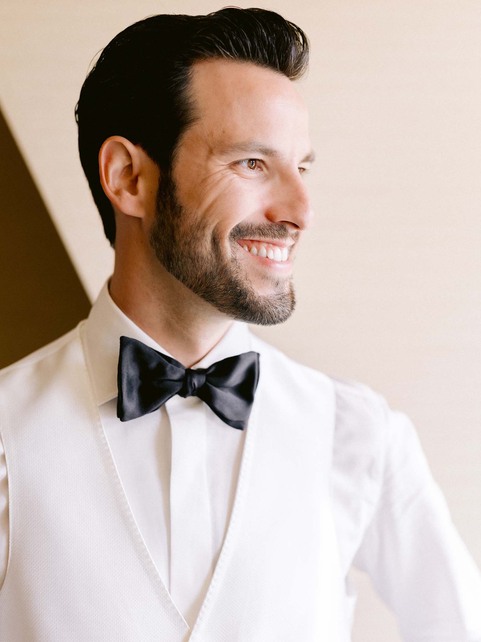 Groom in a white dinner jacket and black bow tie, smiling in the window light