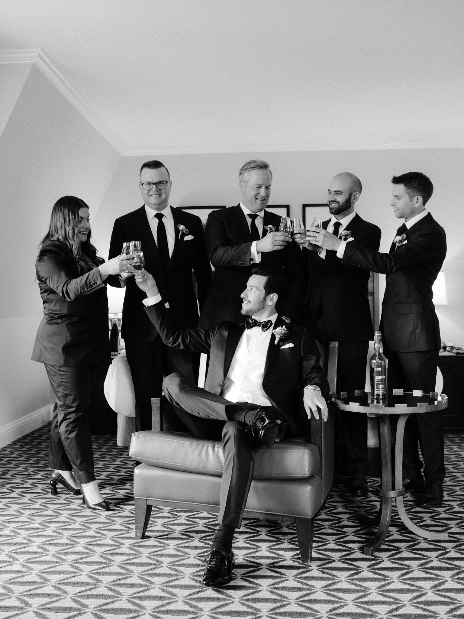 The groom's wedding party raising whisky glasses over his armchair in the hotel suite
