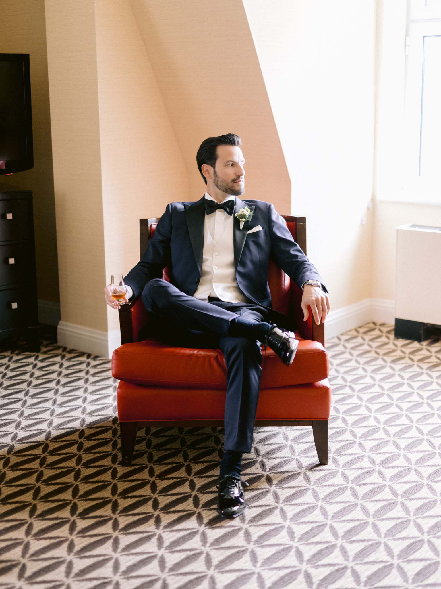 Groom in a navy tuxedo seated in a red leather armchair with a glass of whisky