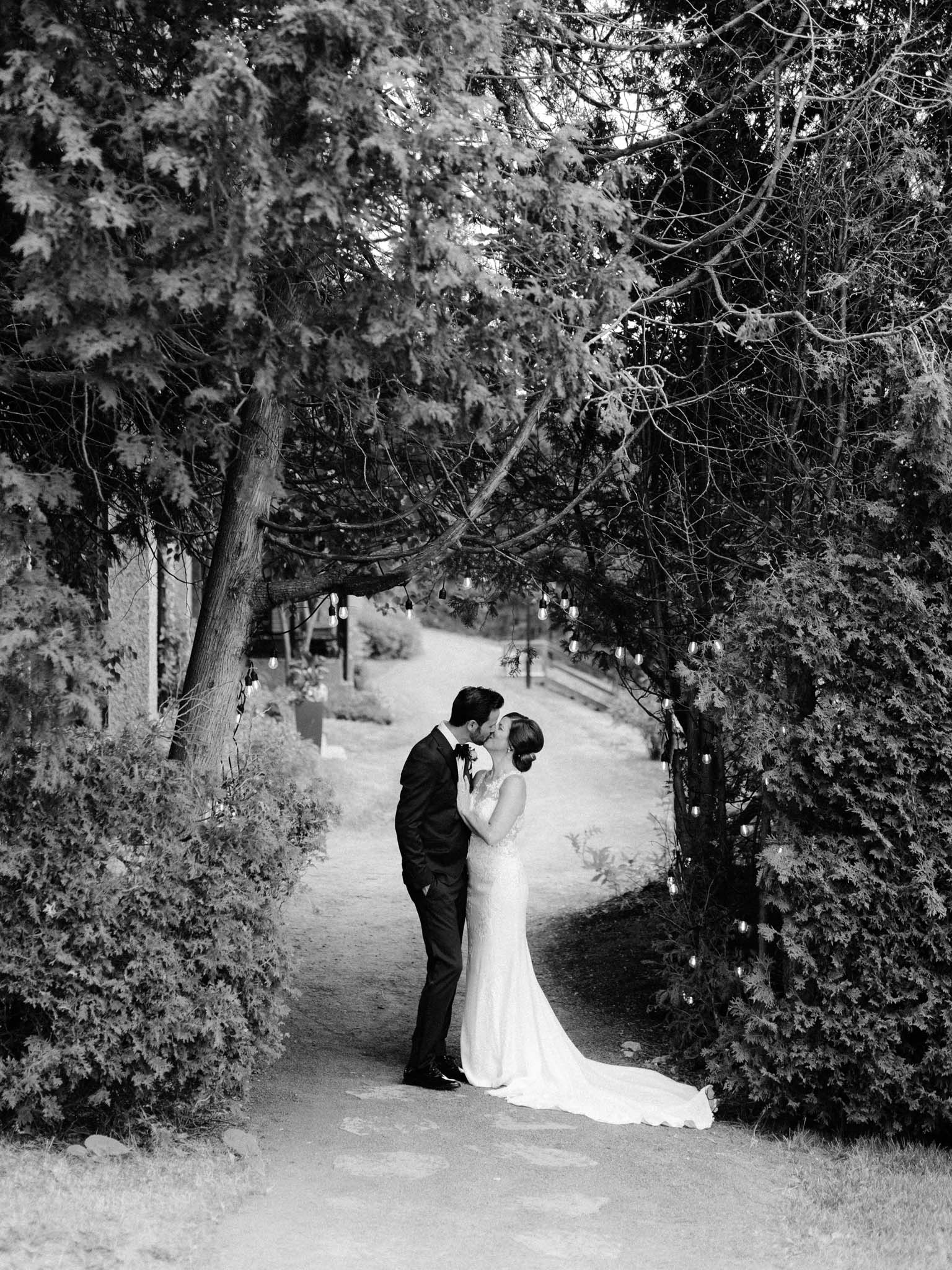 Bride and groom kissing under a canopy of trees strung with bulb lights, in black and white