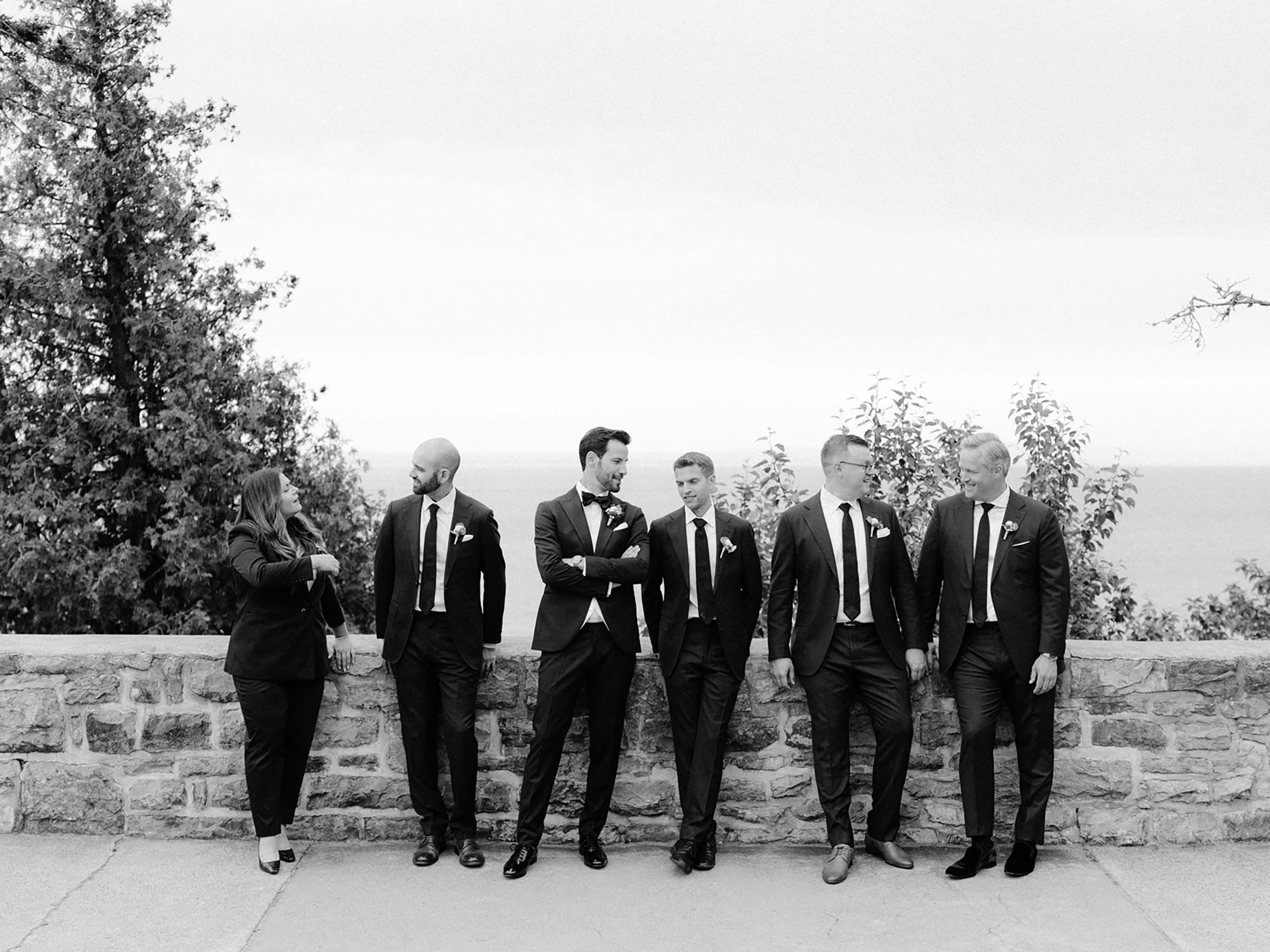 Black and white portrait of the groom and his wedding party leaning against the stone wall above the water