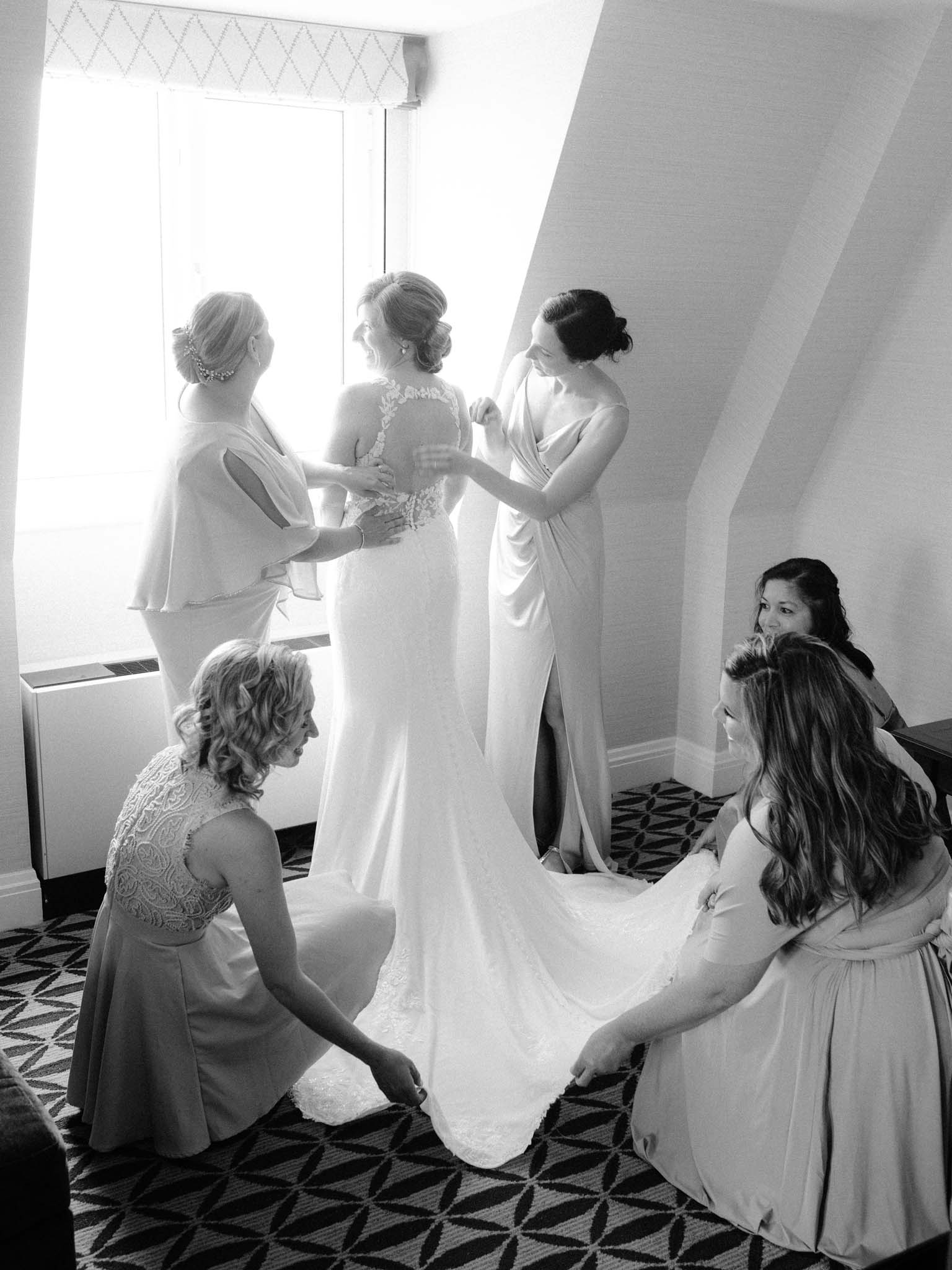 Bridesmaids fastening the bride's lace illusion back and spreading her train by the window