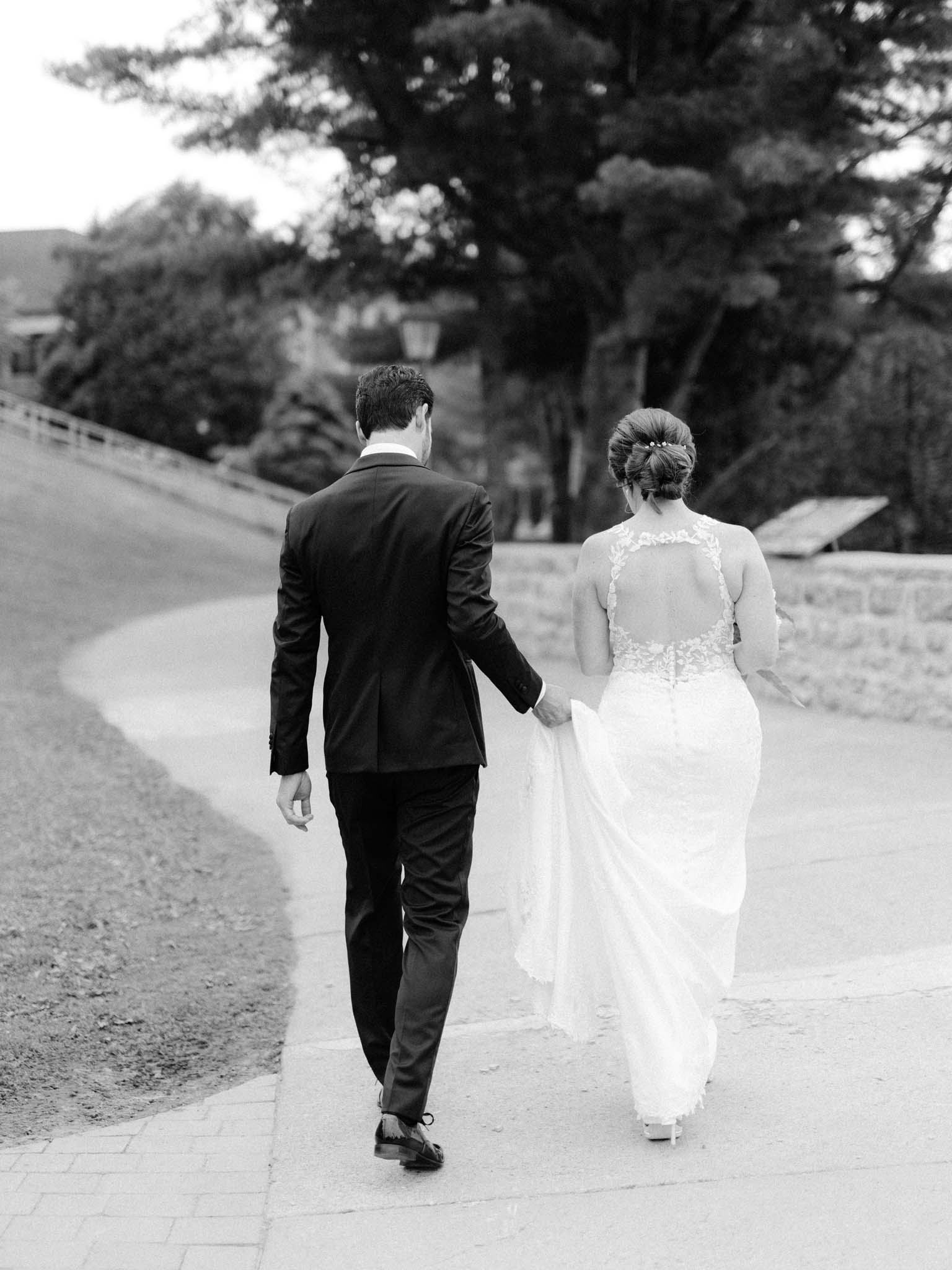 Groom carrying the bride's train as they walk the path together, seen from behind