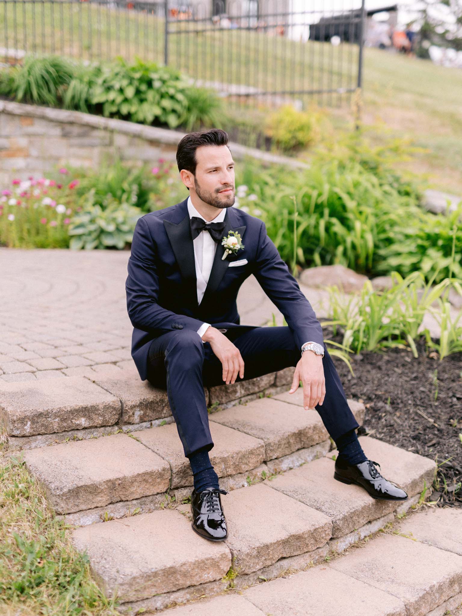 Groom sitting on the stone steps beside the flower beds in his navy tuxedo