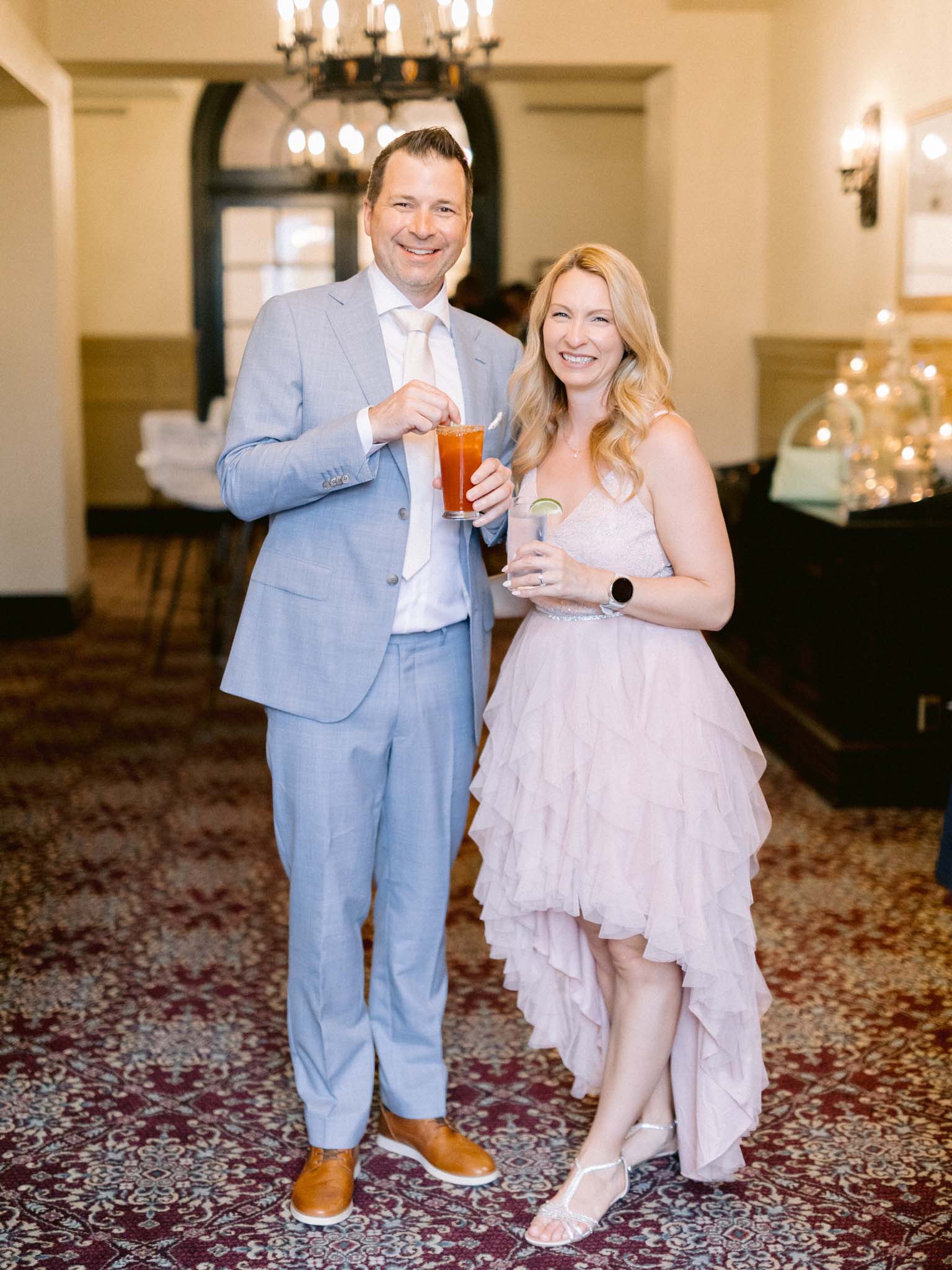 Two guests in a pale blue suit and a blush tulle dress raising their drinks at cocktail hour