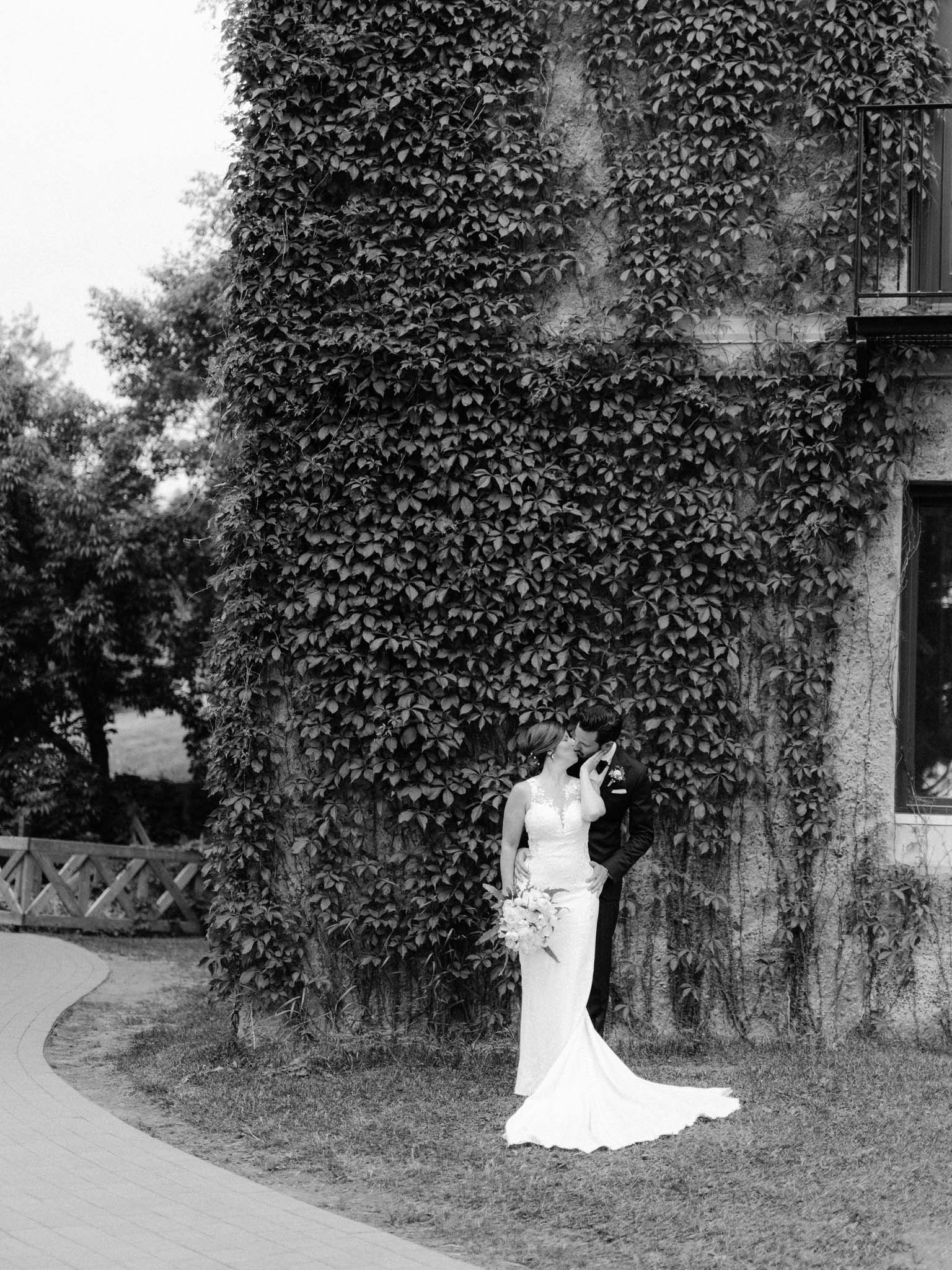 Bride and groom kissing beneath a wall of ivy, in black and white