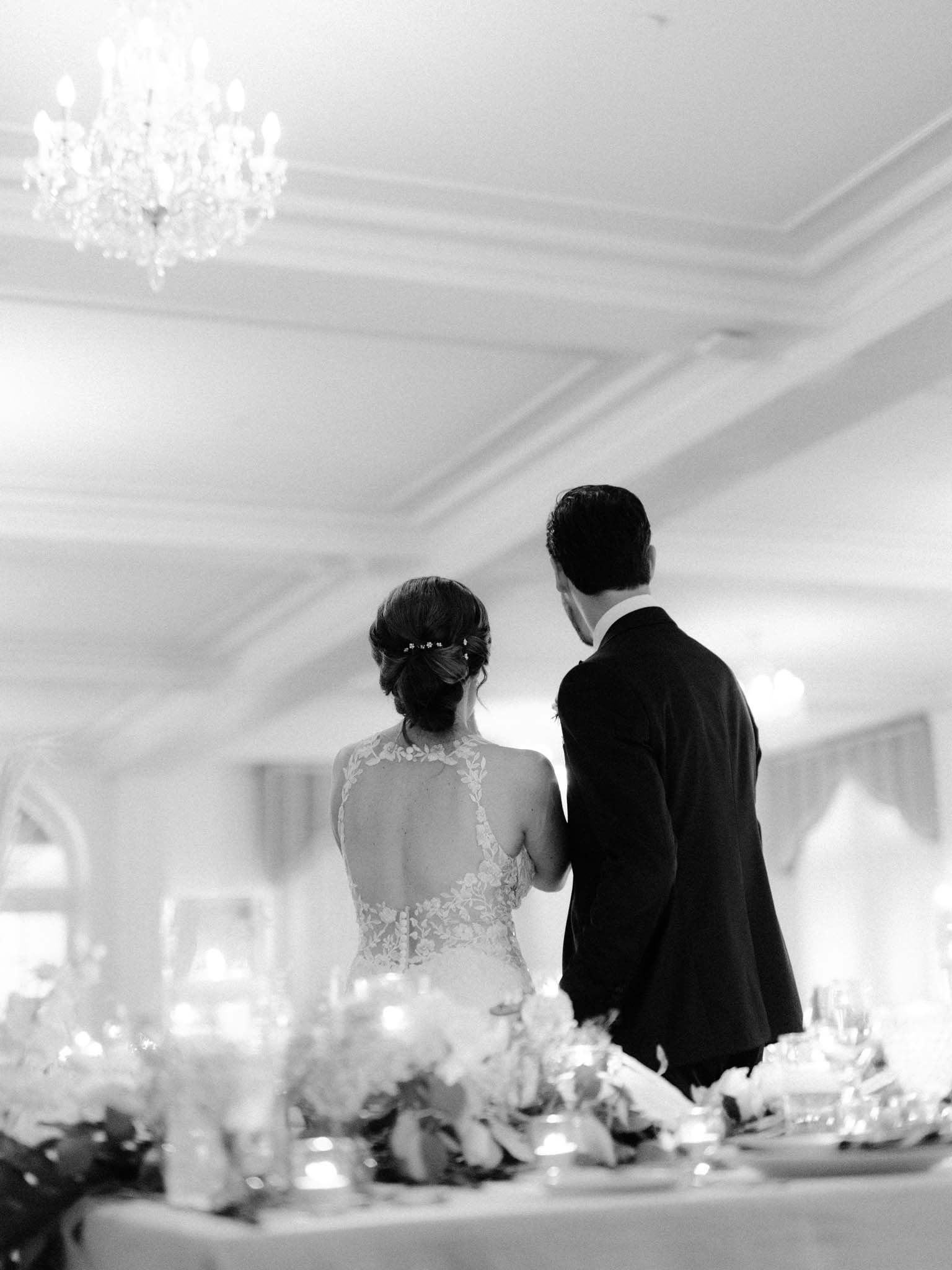 Bride and groom seen from behind as they take their seats at the head table, in black and white