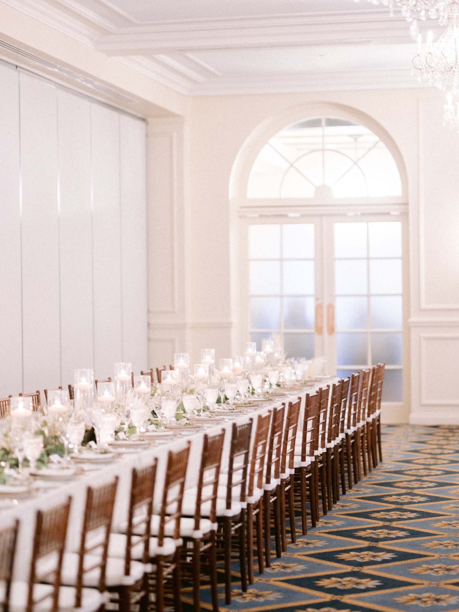 The reception room set with long tables and chiavari chairs beneath the chandeliers