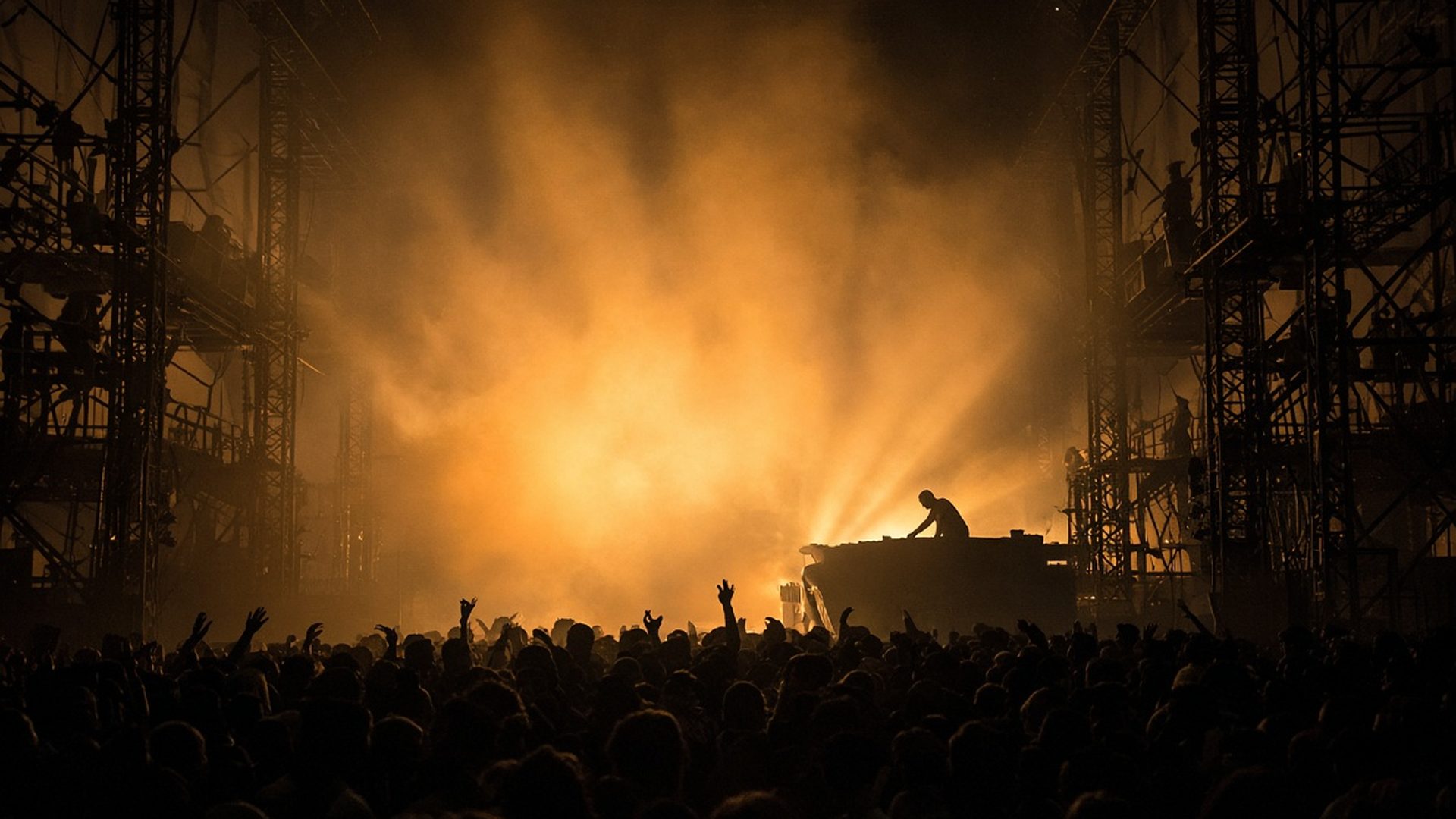 A DJ performs above a crowd with raised hands in orange stage haze