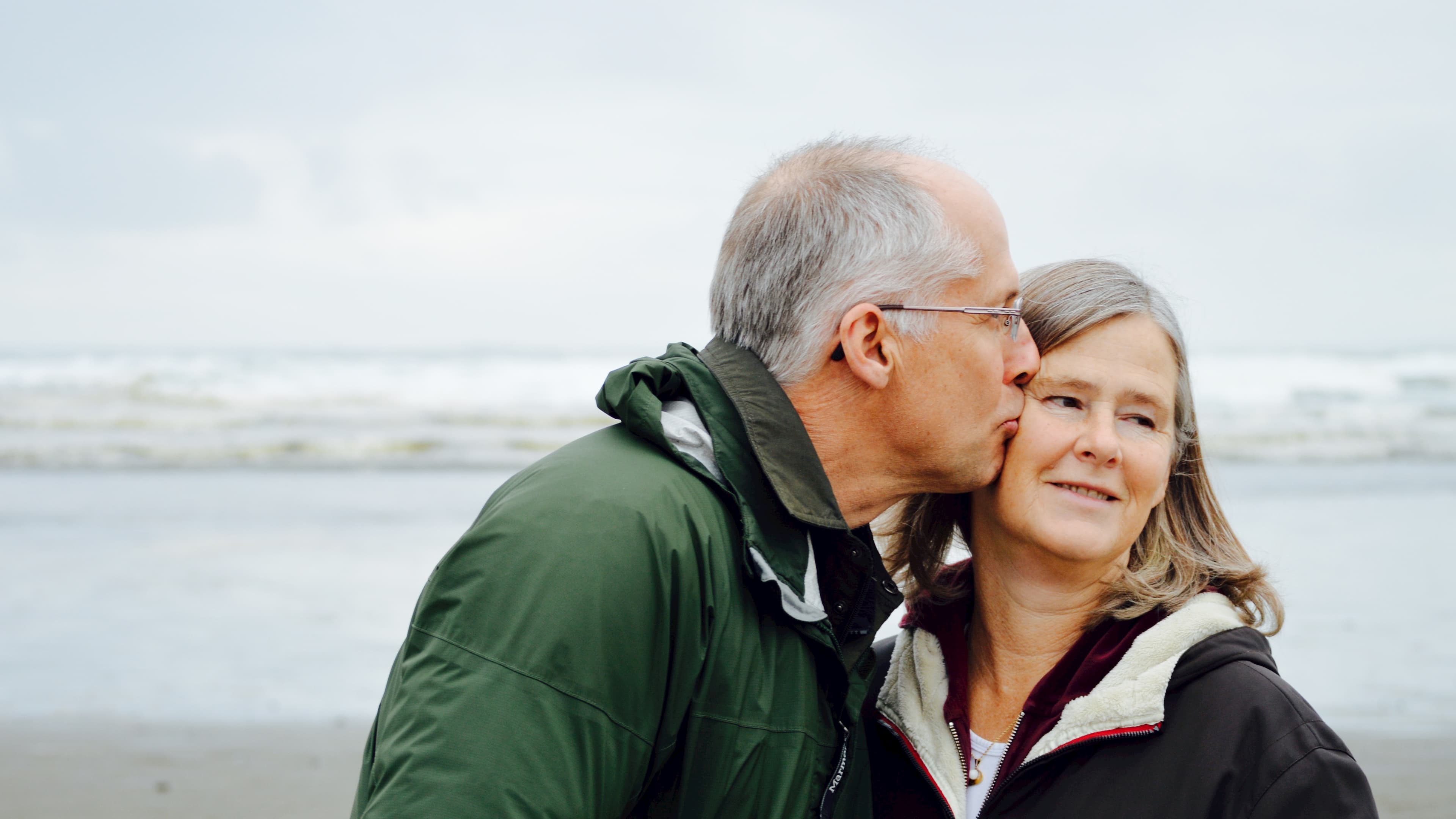 Elderly man kissing elderly woman on cheek in front of beach on cold day