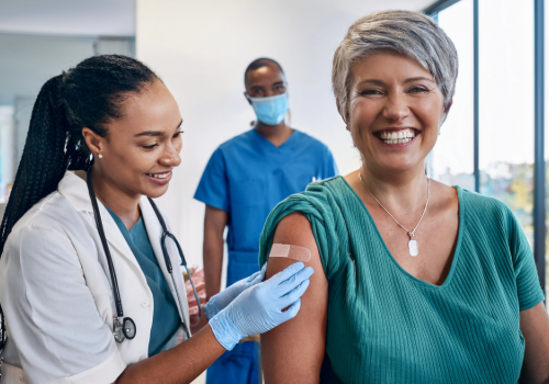 A diverse group of adults (different ages and ethnicities) smiling after getting vaccinated at a clinic, each with a small bandage on their upper arm. Include a nurse or provider in the background giving a flu shot in a clean, friendly environment.