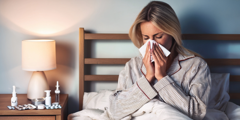 A woman blowing her nose in bed with a thermometer and medications on her nightstand.