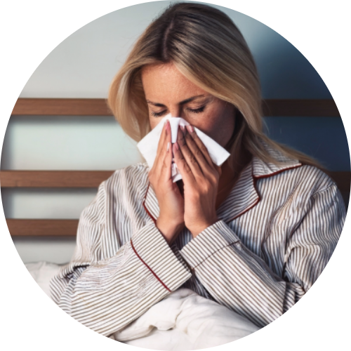 A woman blowing her nose in bed with a thermometer and medications on her nightstand.