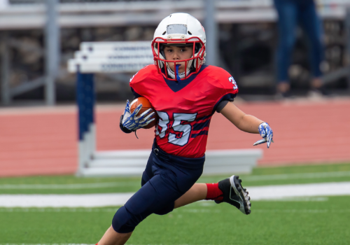 Young kid running with a football in protective gear on a multi-purpose field.