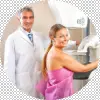 Smiling woman in a pink gown standing beside a male doctor at a mammogram machine, both smiling during a breast screening exam.