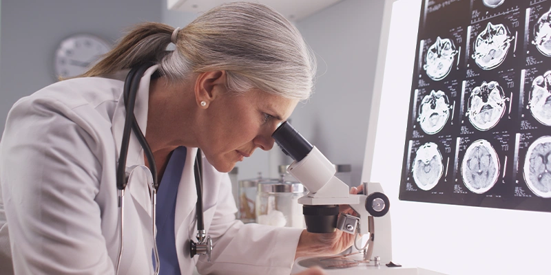 Neurologist reviewing a specimen under a microscope with MRI brain scans beside her.