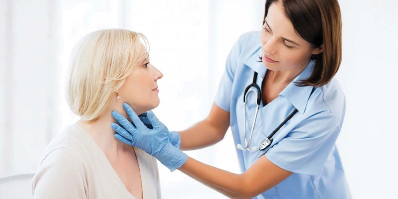 Female ENT doctor with dark hair wearing blue scrubs and gloves gently feeling a blonde female patient’s neck and thyroid area during an exam.