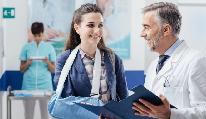 Orthopedic doctor smiling with a patient who has her arm in a sling as they review her chart together, representing compassionate trauma care and recovery after injury.