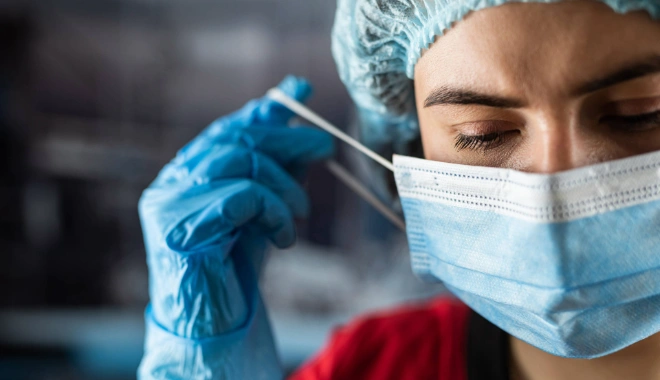 Female nurse wearing protective gear and gloves as she adjusts her medical mask, representing infection control and infectious disease care.