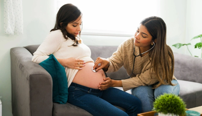 Certified nurse midwife performing an at-home prenatal checkup, listening to a pregnant woman’s abdomen with a stethoscope during a home visit.
