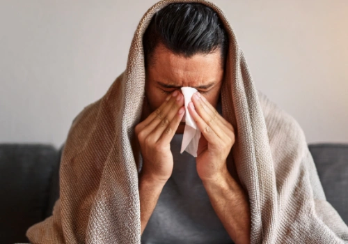 A person holding their forehead and nose bridge with a tissue in hand, sitting on a couch under a blanket, showing discomfort. Natural lighting with soft focus on the sinuses area.